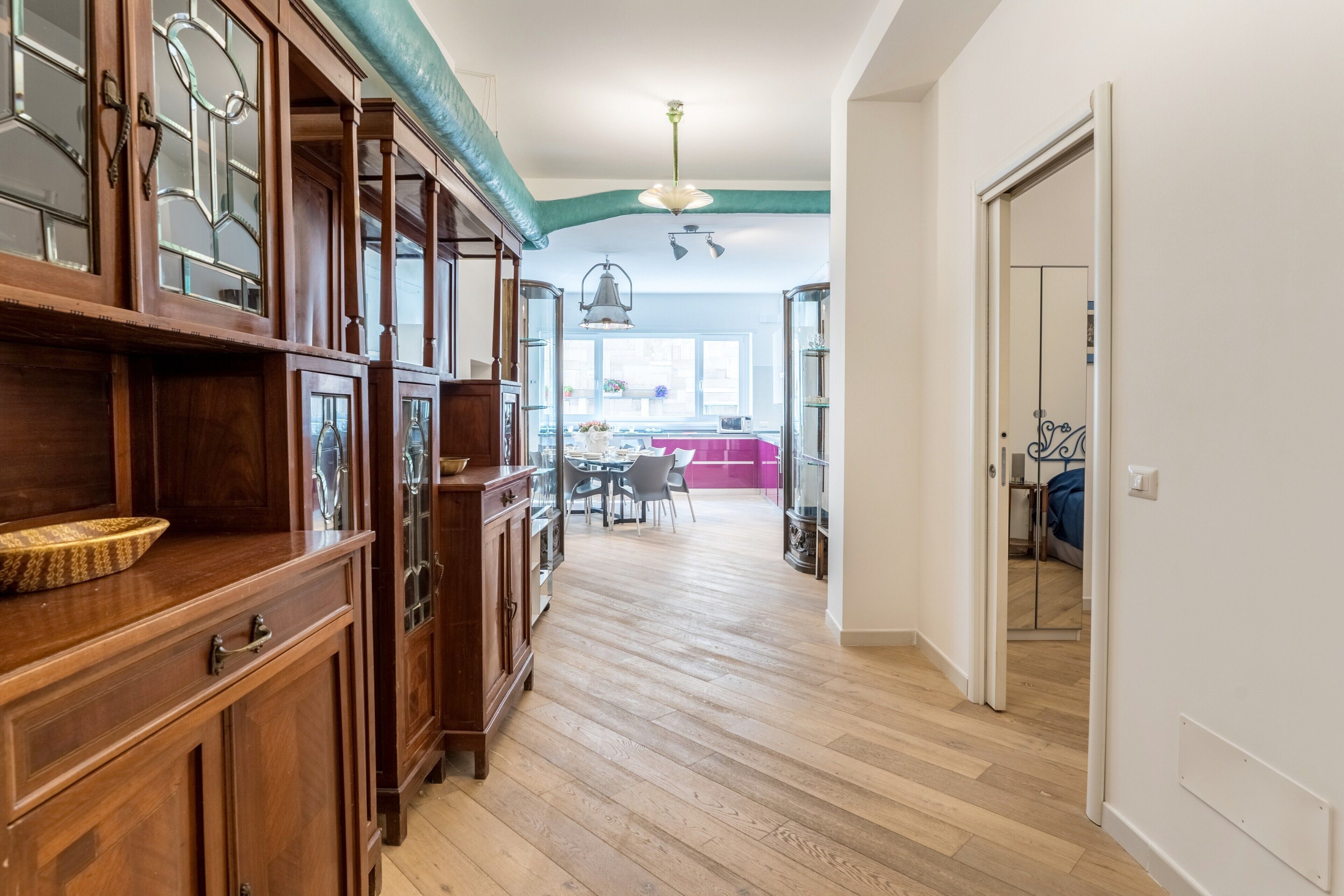 A hallway with wooden cabinets, a dining area with a pink table, and a kitchen with a window.