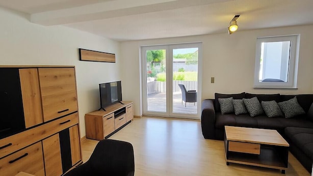 Una sala moderna con una televisión de pantalla plana, muebles de madera, un sofá con cojines estampados y un amplio ventanal corredizo que da a un patio exterior.