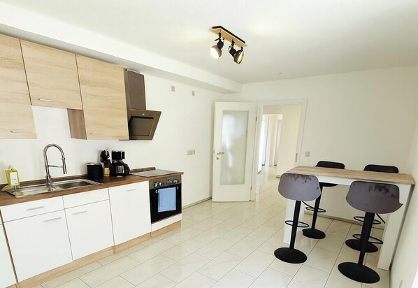 A modern kitchen with white cabinets, a built-in oven, and a coffee maker. There is a dining area with a table and four bar stools.