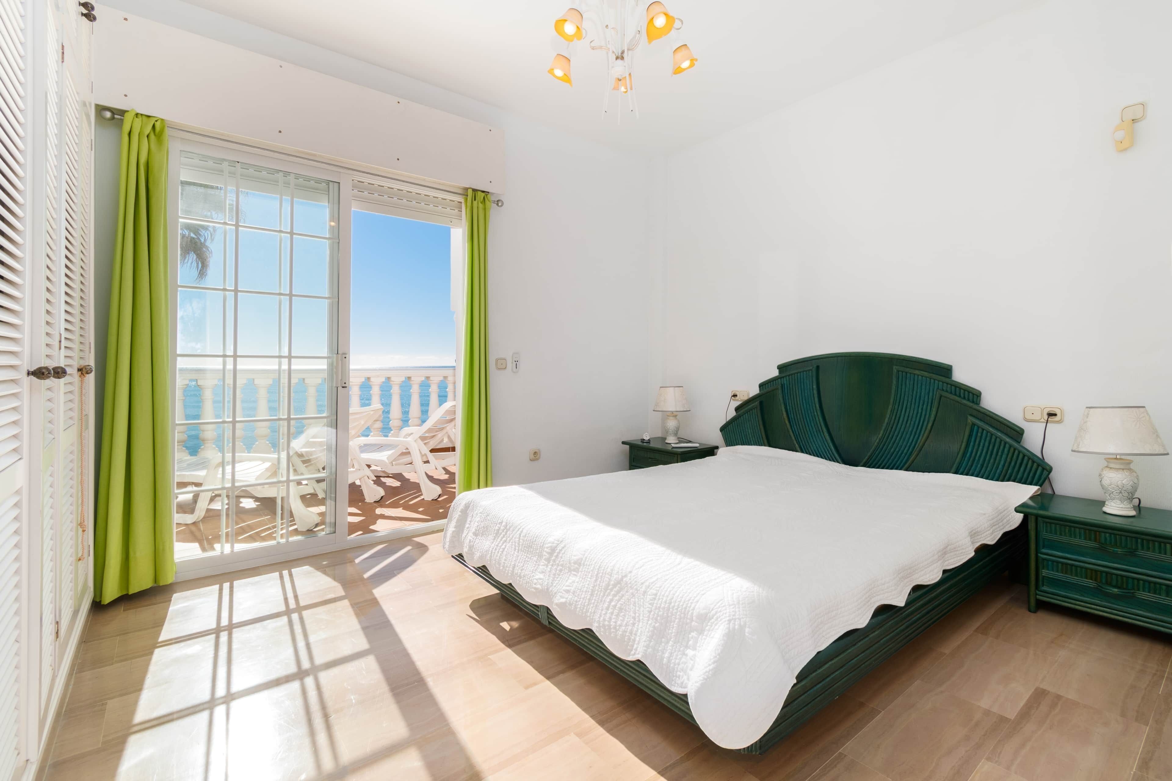 A bedroom with a bed, a nightstand, a lamp, a window with curtains, and a view of the outside.