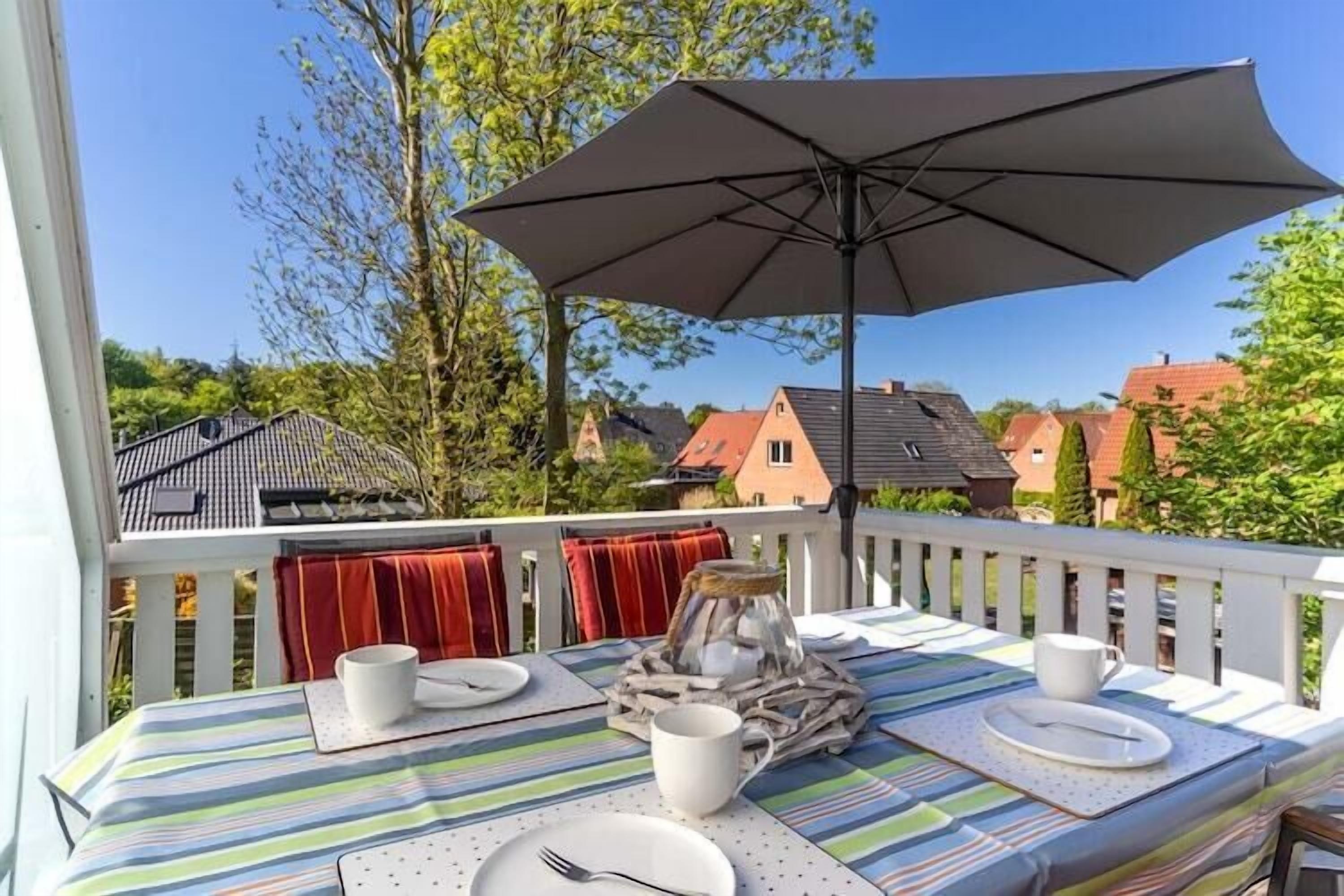 Un balcón con mesa para dos, un gran paraguas y vista a casas y árboles.