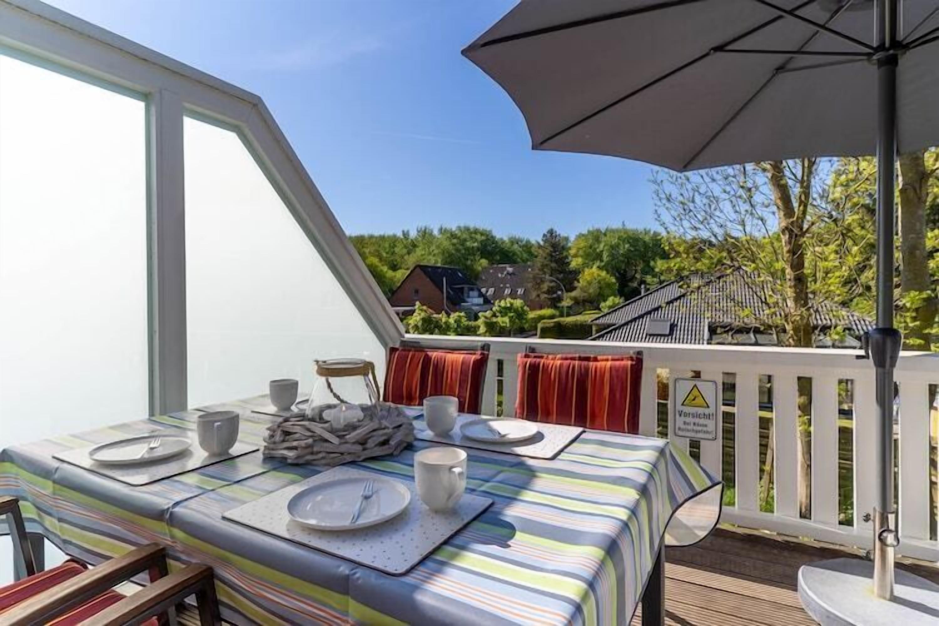 A dining area with a table set for two, a large umbrella, and a view of a house and trees.
