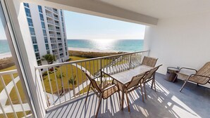 Un balcon avec une table et des chaises en verre, donnant sur une plage et un immeuble de grande hauteur.