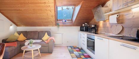 A cozy loft-style room with a wooden ceiling, a small window, a sofa with cushions, a round coffee table, a kitchen area with white cabinets and a stove, and a colorful rug.