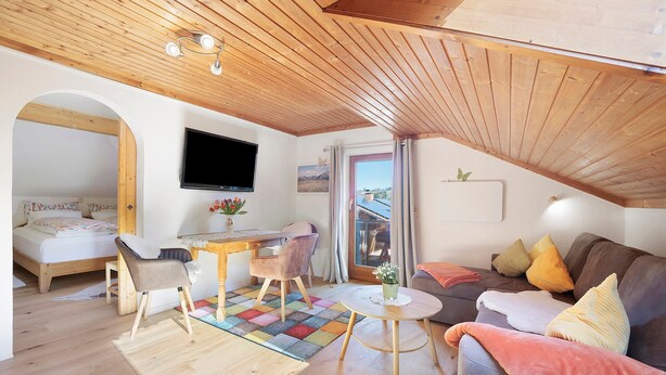 A cozy room with a bed, a TV, a dining area, and a couch with pillows. - Apartment 'Haus Bergliebe – Zugspitzblick' with Mountain View, Balcony and Wi-Fi (Pfronten)