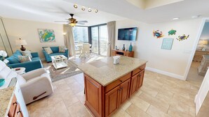 A spacious living area with a blue sofa, a coffee table, and a TV. There is a kitchen with wooden cabinets and a granite countertop. - Mainsail 223 Stunning Gulf Views & Beach Service (Miramar Beach)