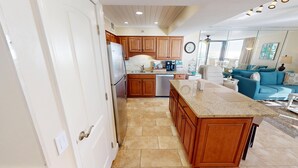 A kitchen with wooden cabinets, a built-in island, and a dining area with a blue sofa and a coffee table. - Mainsail 223 Stunning Gulf Views & Beach Service (Miramar Beach)