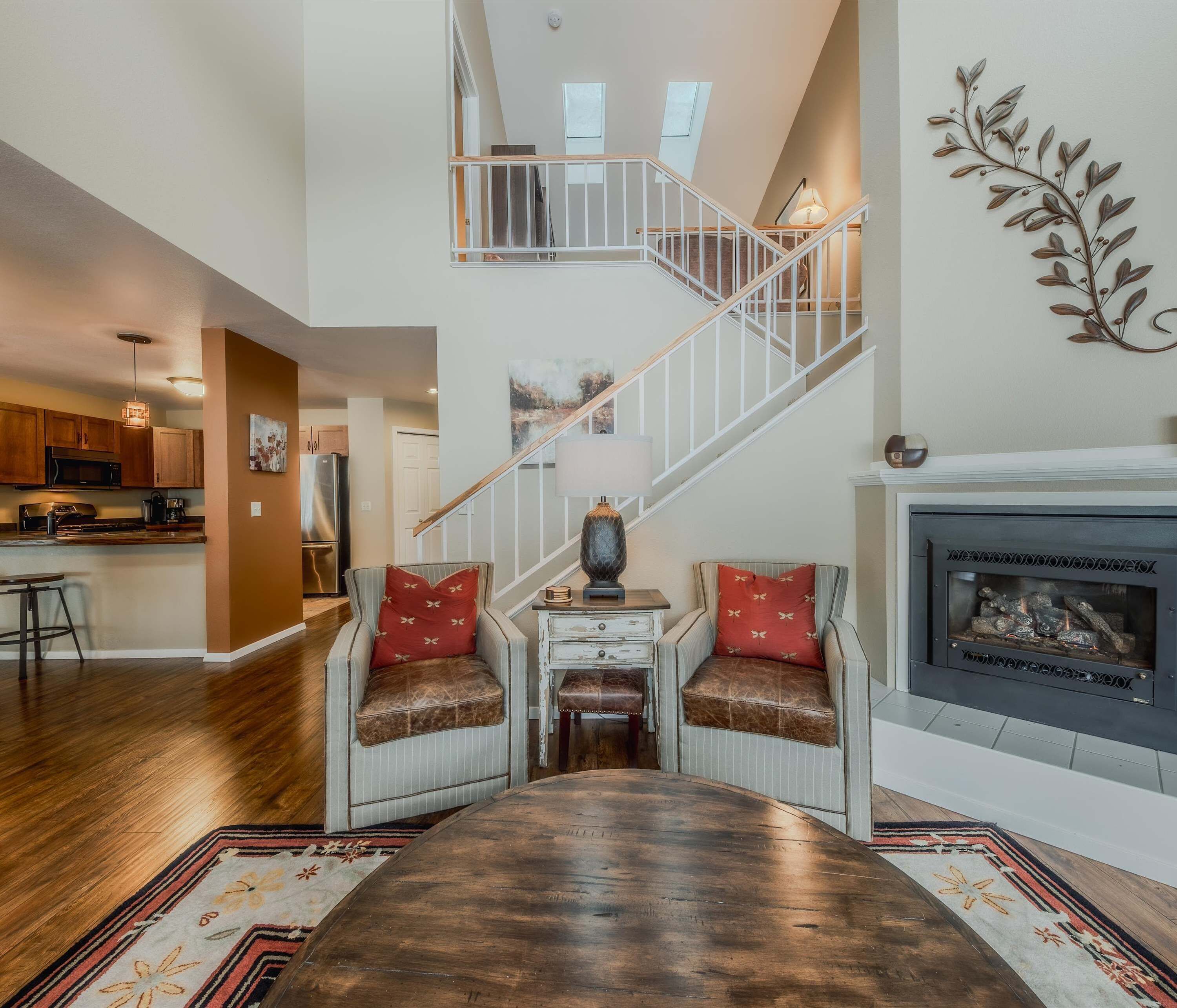 A spacious living area with a wooden floor, a fireplace, a staircase, and a kitchen in the background.