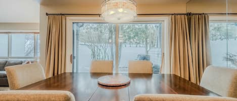 Un coin repas avec une table en bois et quatre chaises, un luminaire suspendu et de grandes fenêtres avec des rideaux.