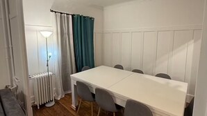Un coin repas avec une table blanche et des chaises grises, un radiateur et une fenêtre avec des rideaux.