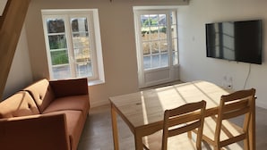 A room with a sofa, a dining table with chairs, a TV mounted on the wall, and two windows with a view of the outside. - Gîte de Vouhé near Surgères apartment 2 (Vouhé)