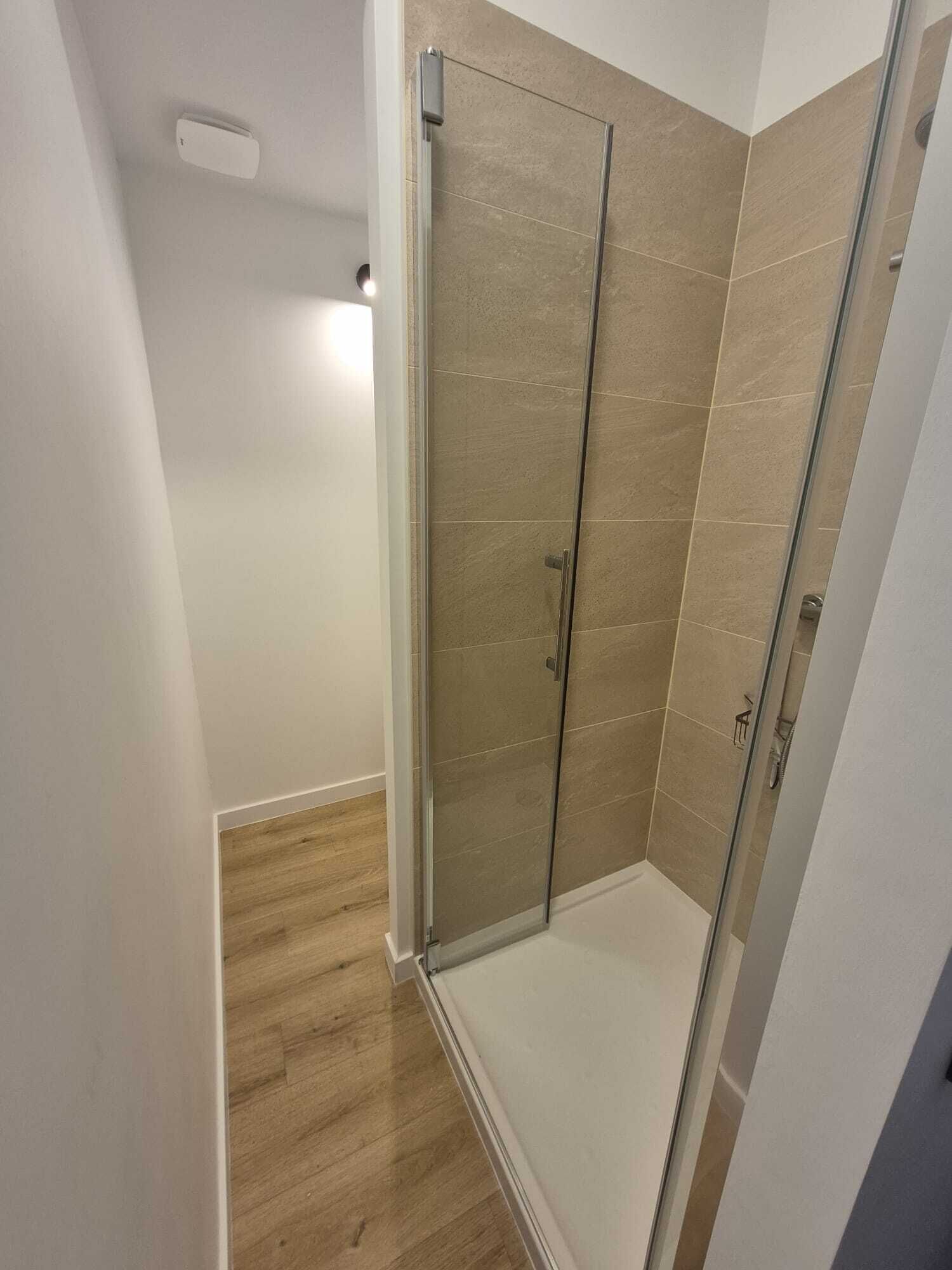 A modern bathroom with a glass shower enclosure, wooden flooring, and beige tiles.