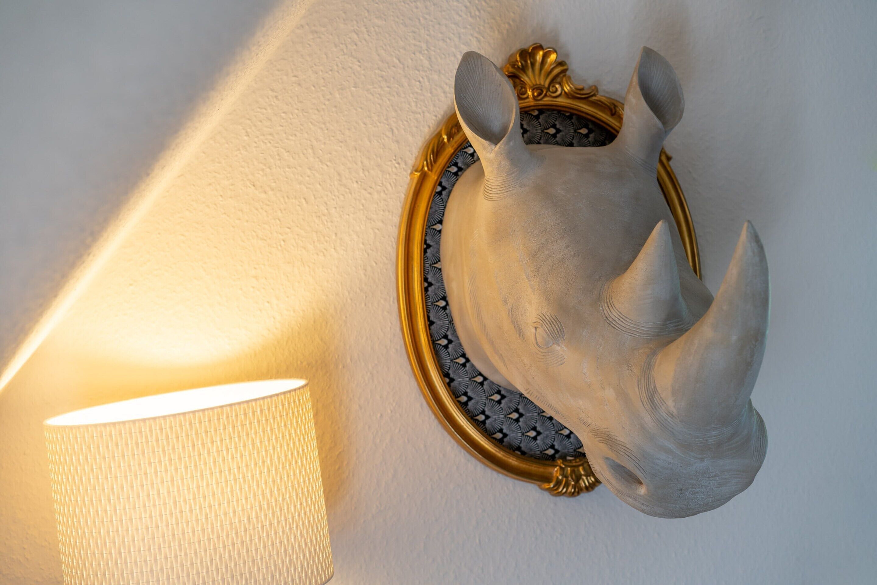 Une sculpture murale représentant la tête d’un rhinocéros, encadrée d’un cadre doré, placée à côté d’une lampe fixée au mur.