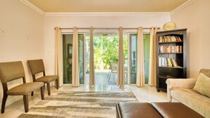 Ein Wohnzimmer mit einer Couch, Sesseln, einem Bücherregal und Blick durch Glastüren auf einen Außenbereich.