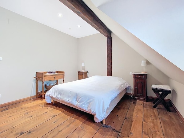 A bedroom with a wooden floor, a bed with white bedding, a wooden nightstand, a small table, and a lamp.
