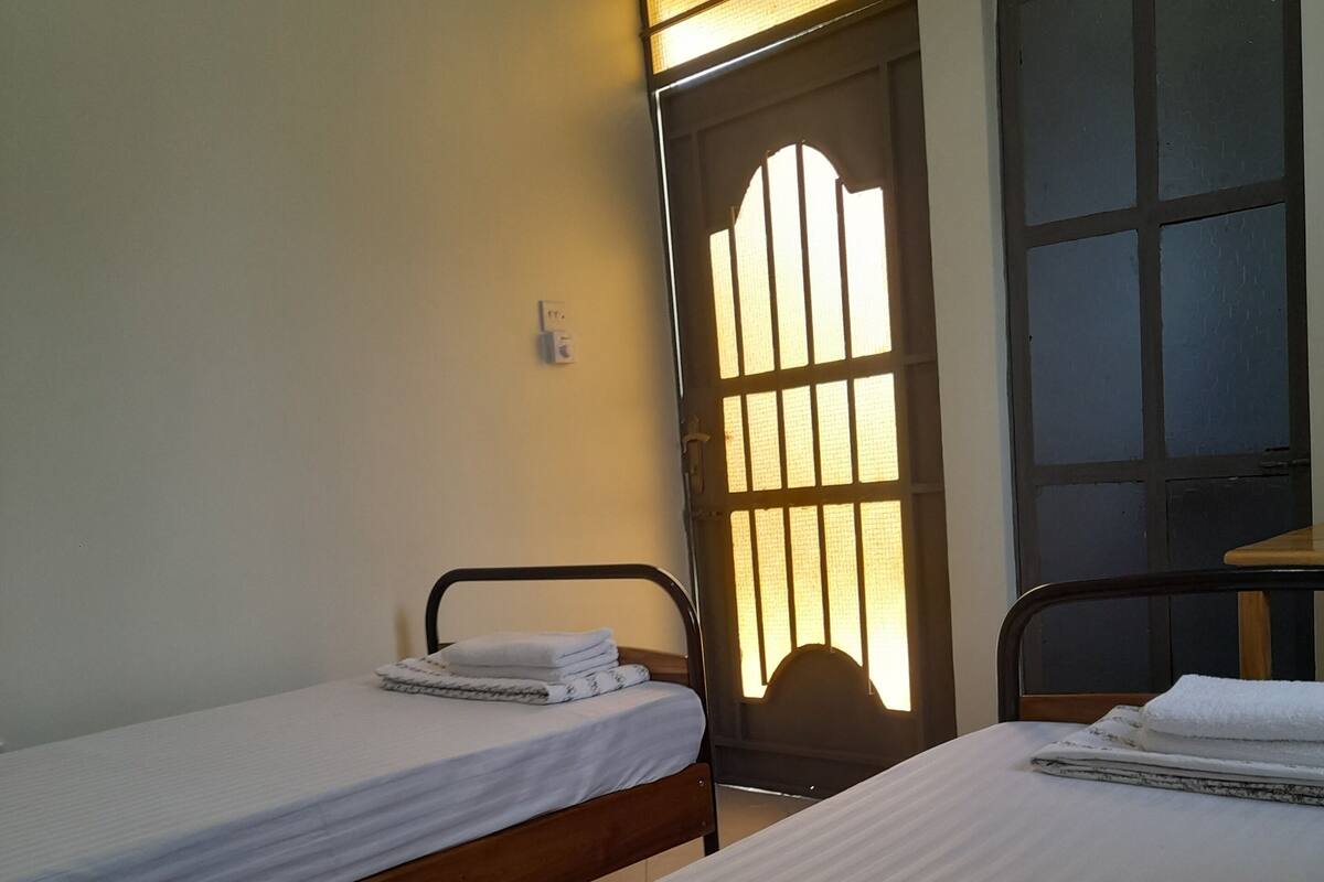 A room with two single beds, a wooden headboard, a small table, and a window with a view of the outside.