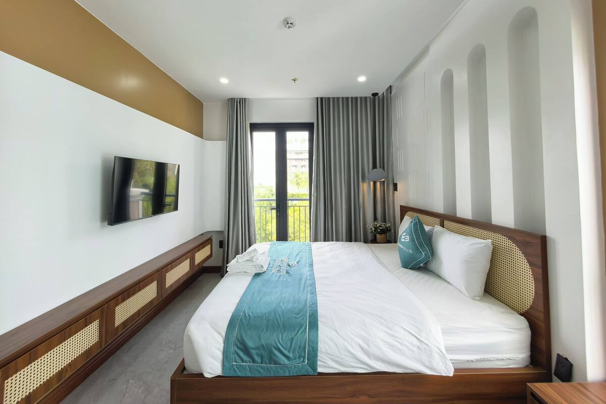 A hotel room with a large bed, a flat-screen TV mounted on the wall, and a balcony with curtains.