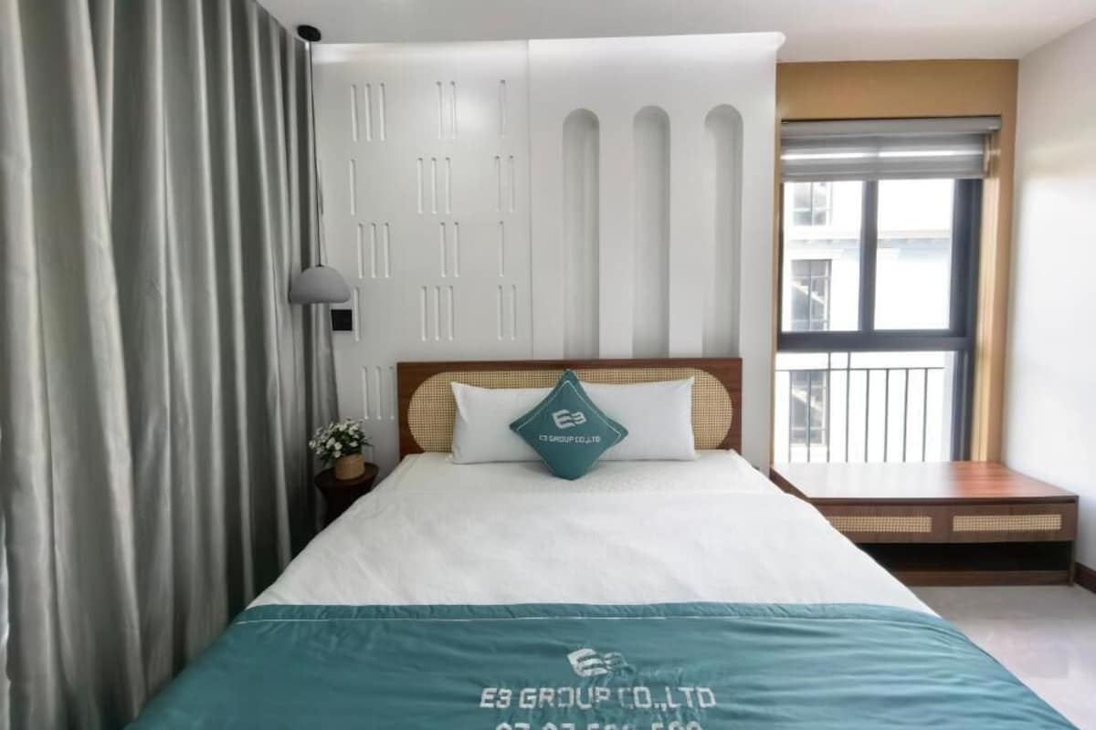 A hotel room with a bed, a nightstand, a lamp, a window with blinds, and a view of the outside.