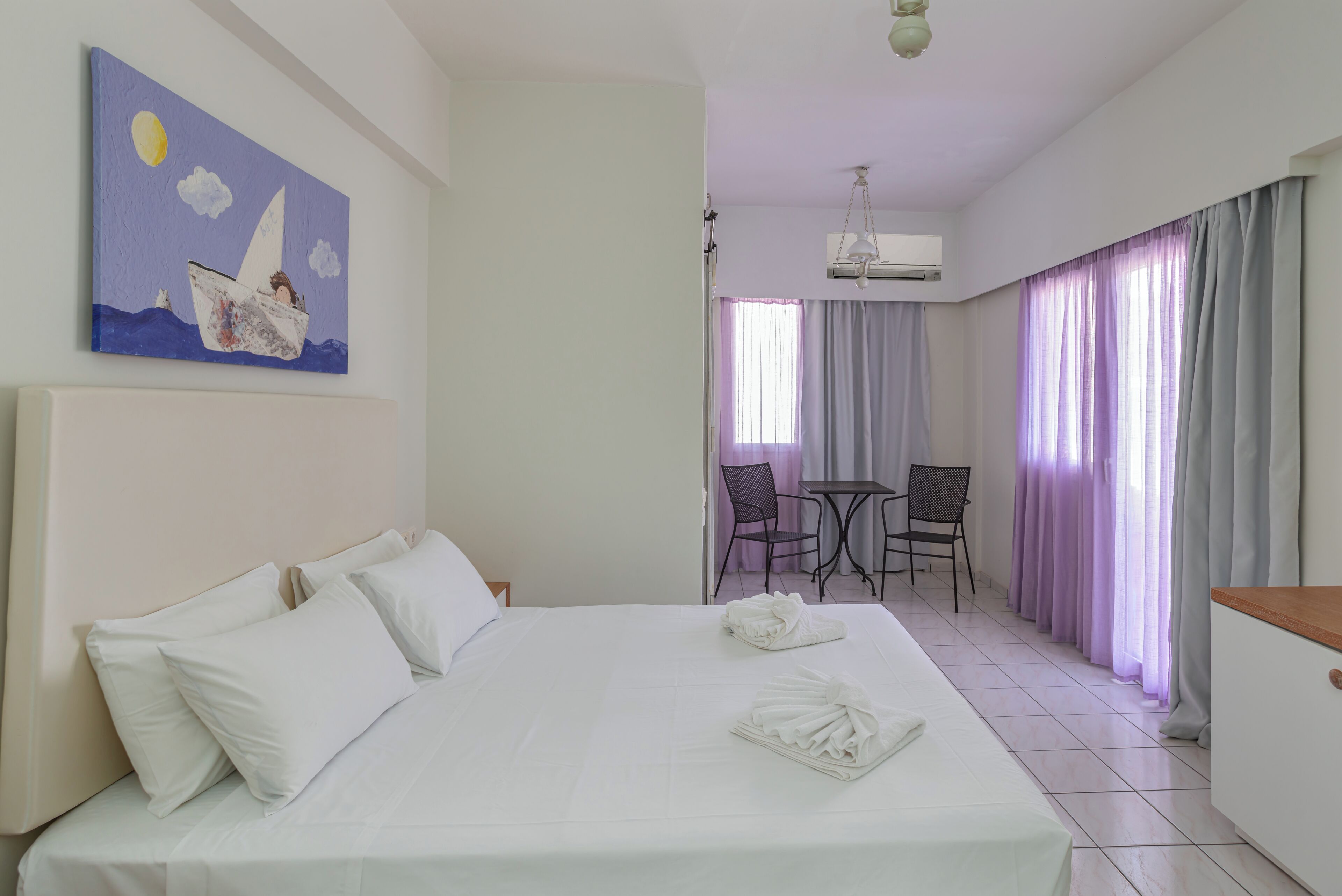A hotel room with a bed, pillows, a painting, a dining area with chairs and a table, and a window with curtains.