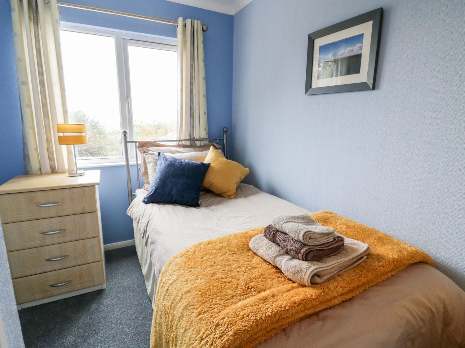 A bedroom with a bed, a bedside table, a lamp, a window with curtains, and a framed picture on the wall.
