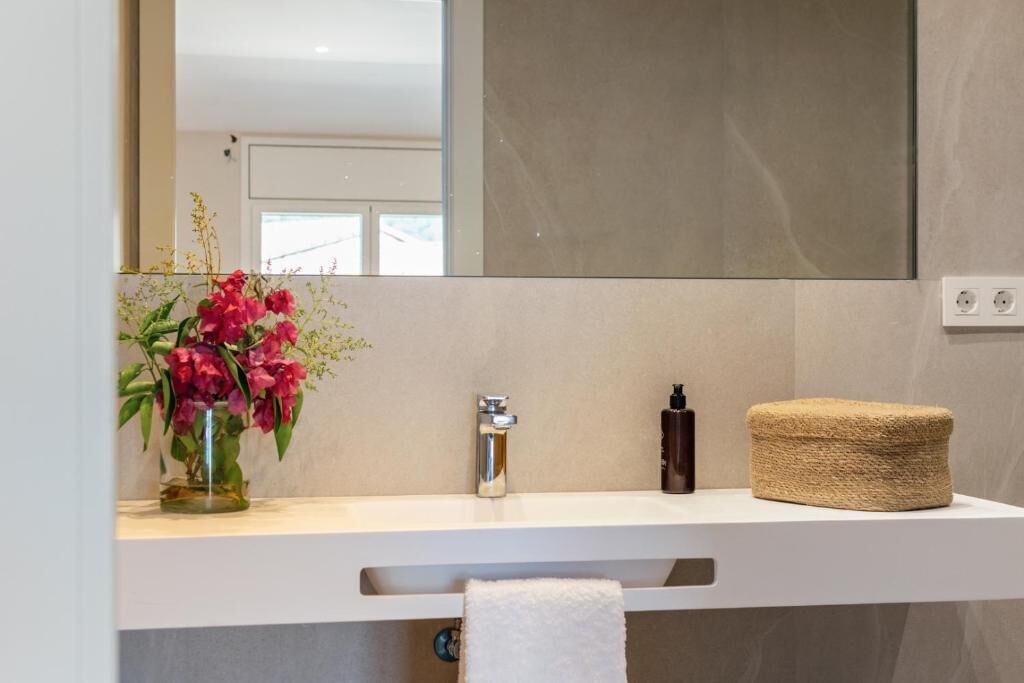 Un baño con tocador blanco, espejo, un jarrón de flores, toalla y botella de champú.