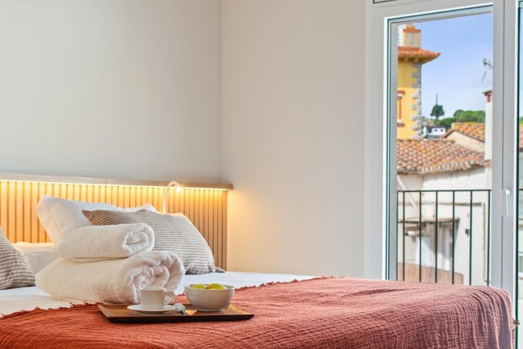 A hotel room with a bed, towels, a tray with a cup and bowl, and a view of buildings through a balcony.