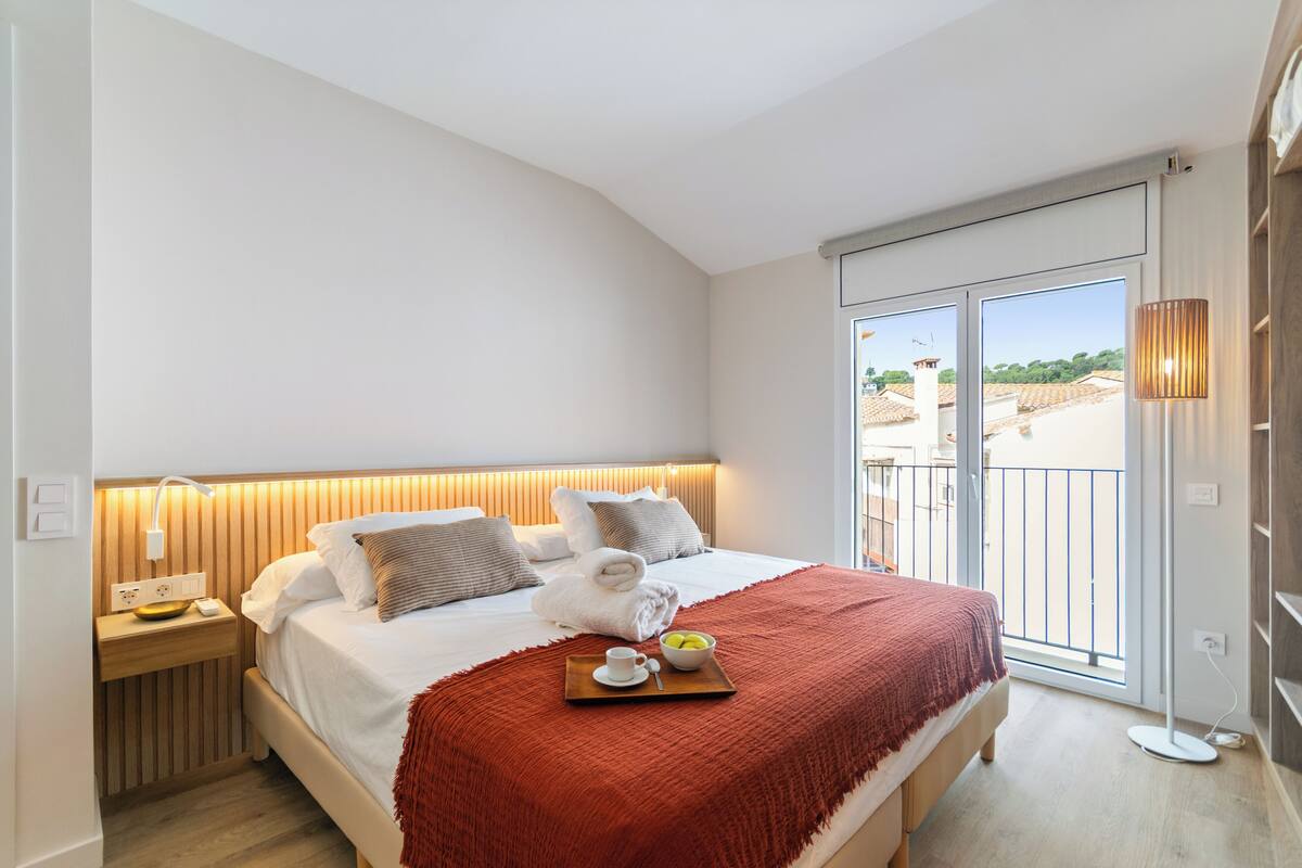A hotel room with a bed, a bedside table with a lamp, a tray with a cup and fruit, and a view of the outside.