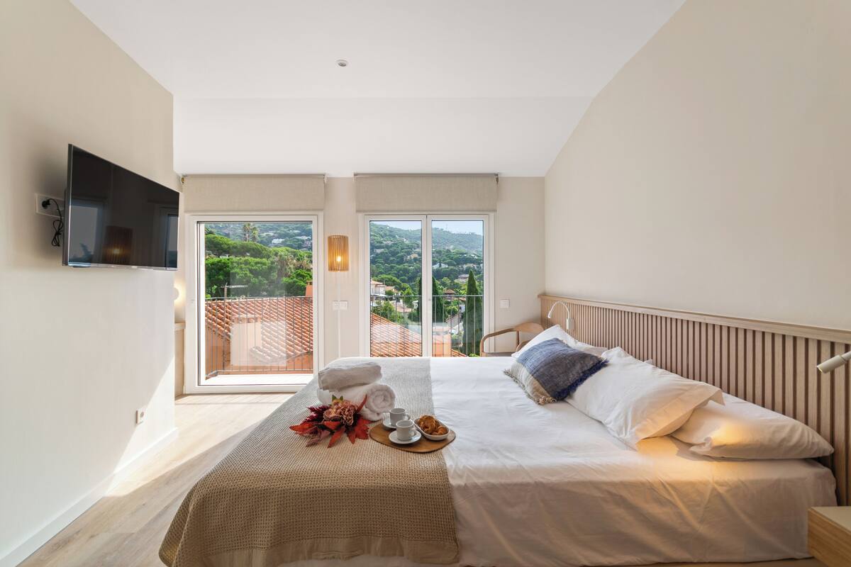 A bedroom with a bed, a TV mounted on the wall, and a balcony with a view of greenery.