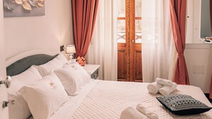 Un letto rifatto con lenzuola bianche e un motivo trapuntato, un comodino con una lampada e una finestra con tende rosse.