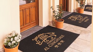 Ingresso di un hotel con porte in legno, piante in vaso e tappetini neri con scritte.
