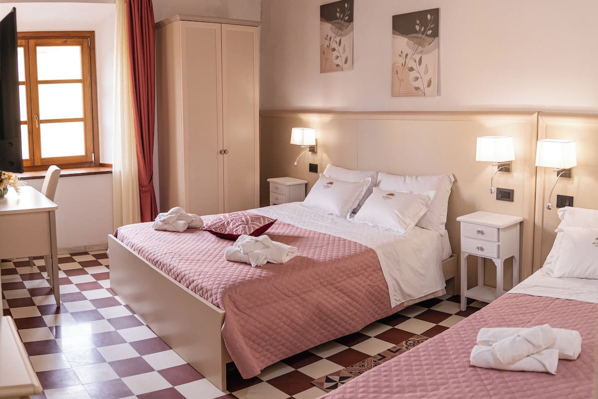 A hotel room with a checkered floor, a bed with pink bedding, a desk with a chair, a wardrobe, and two wall-mounted lamps.