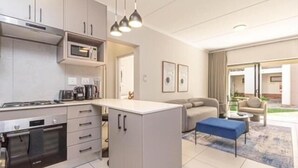 A modern kitchen with a built-in microwave, oven, and coffee maker. There is a small island with a sink and a blue bench. The room has a neutral color palette and a large window. - The Henlee Apartment Dainfern (Dainfern)