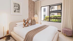 A neatly arranged hotel room with a large bed, a side table, a lamp, and a window with a view of greenery. - The Henlee Apartment Dainfern (Dainfern)