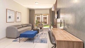 A modern living room with a sofa, a coffee table, and a wooden side table. There are two framed artworks on the wall and a large window with curtains. - The Henlee Apartment Dainfern (Dainfern)