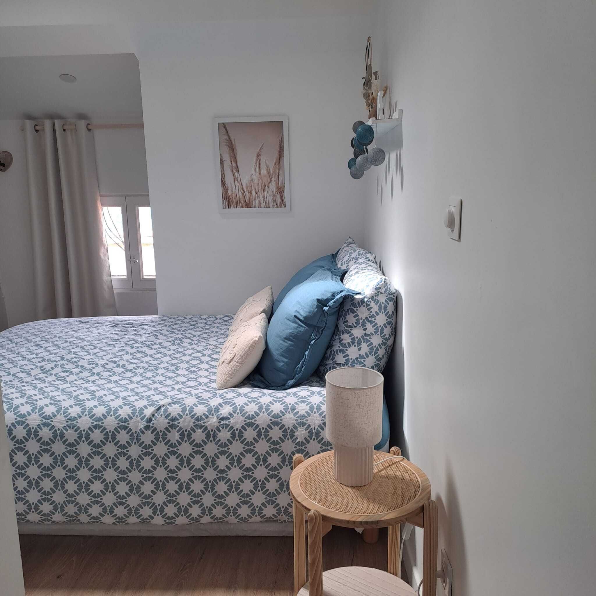 A bedroom with a bed, pillows, a small round table with a lamp, and a wall-mounted shelf with decorative items.