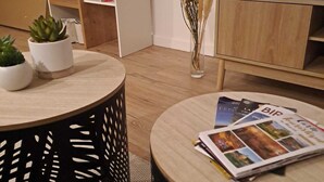 Ein modernes Wohnzimmer mit einem runden Holztisch, einem schwarzen, gelochten Stuhl, einem kleinen Topfpflänzchen, einer Vase mit Pampasgras und einem Fernseher auf einer Holzkommode.