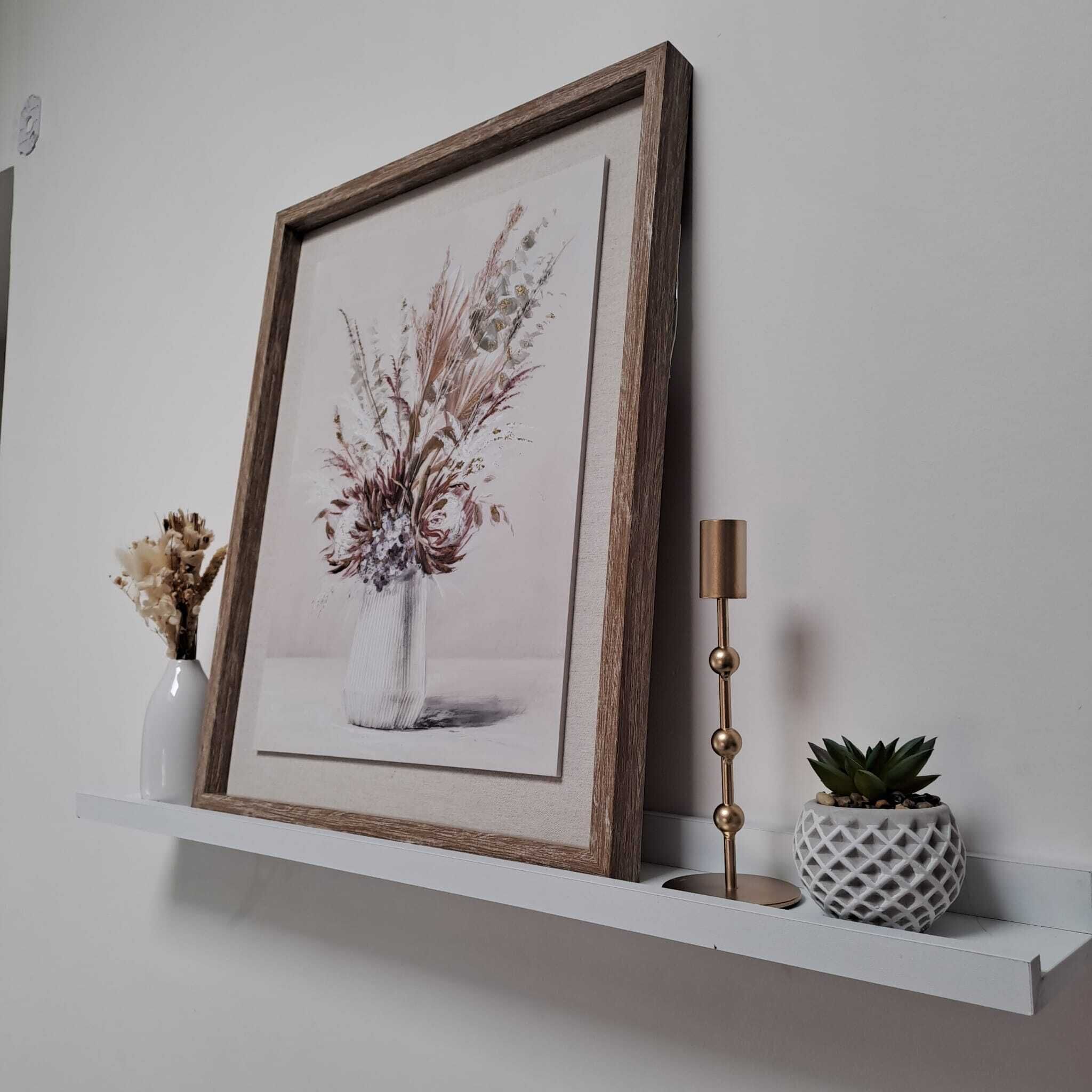 Une œuvre d’art encadrée représentant un vase de fleurs posé sur une étagère blanche, un petit vase contenant des fleurs séchées et un grand chandelier.