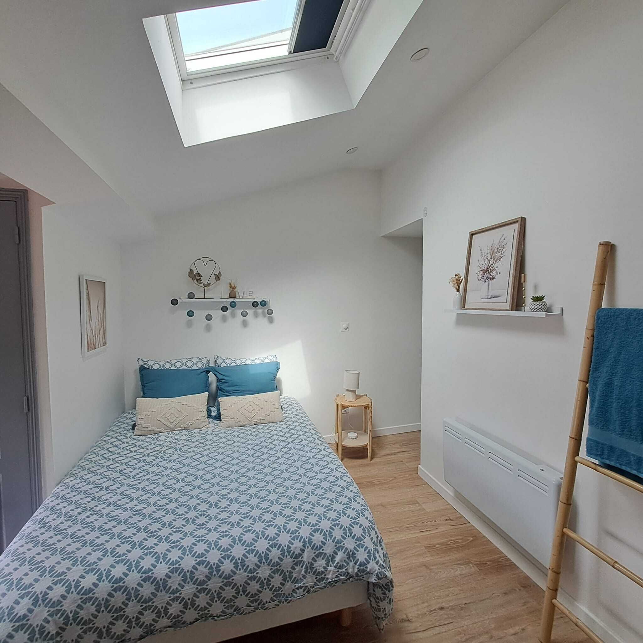 A bedroom with a skylight, a bed with patterned bedding, a bedside table with a lamp, a framed picture on the wall, and a blue towel on a rack.