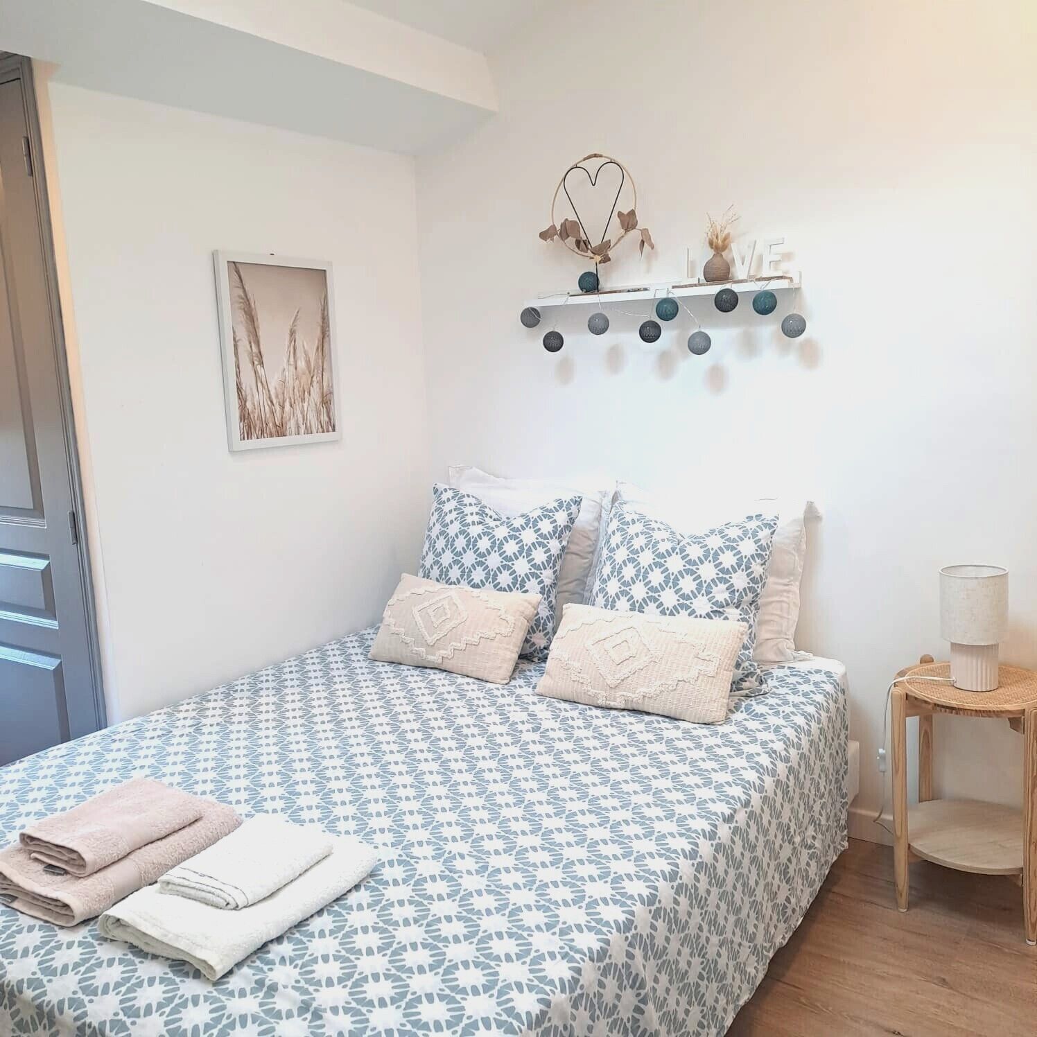 Un dormitorio con una colcha estampada, almohadas, una mesita de noche con lámpara, un estante en la pared con objetos decorativos y un cuadro enmarcado.