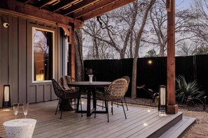Een terras buiten met een houten tafel en rieten stoelen, een kleine witte tafel met twee glazen, en een lantaarnlamp.