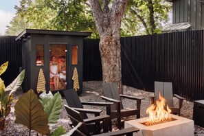 Ein Garten mit einer kleinen Holzkonstruktion, Sitzgelegenheiten im Freien, einer Feuerstelle und einem Baum.