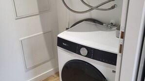 En kompakt, vit tvättmaskin med frontmatning, försedd med en kontrollpanel och slangar, installerad i ett litet tvättrum med trägolv och en vit vägg.