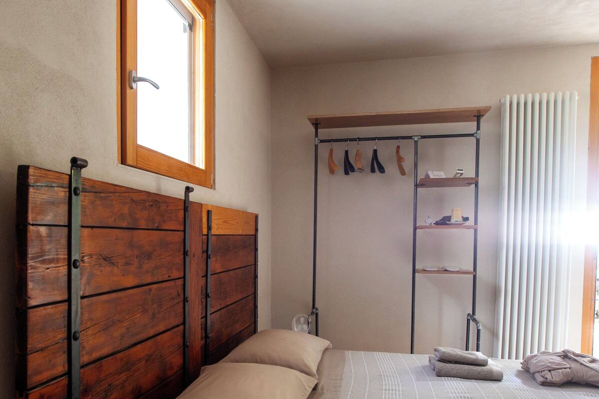 A bedroom with a wooden headboard, a window, a radiator, and a shelf with towels.
