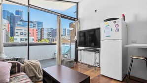 A modern living room with a large window, a flat-screen TV, a refrigerator, and a dining area. - Melb suite zen garden A0 F1 (West Melbourne)