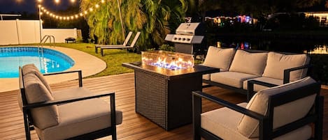 An outdoor deck with a pool, lounge chairs, a fire pit, and a grill area.