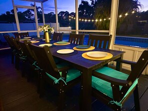 An outdoor dining area with a wooden table and chairs, a vase with flowers, and string lights. - Heated Pool • Fire Pit • 10 Min to Madeira Beach & John`s Pass • Sleeps 10 (St. Petersburg)