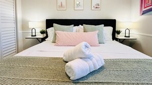 Uma cama bem arrumada com uma toalha enrolada, duas mesas de cabeceira com abajures e três quadros na parede.