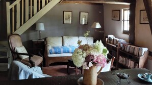 Una sala de estar acogedora con una escalera de madera, un sofá con una manta azul y blanca a rayas, una mesa de centro de madera con un jarrón de flores y una estantería llena de libros.