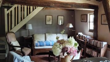 Un salon chaleureux avec un escalier en bois, un canapé recouvert d’un plaid à rayures bleues et blanches, une table basse en bois ornée d’un vase de fleurs et une bibliothèque remplie de livres.