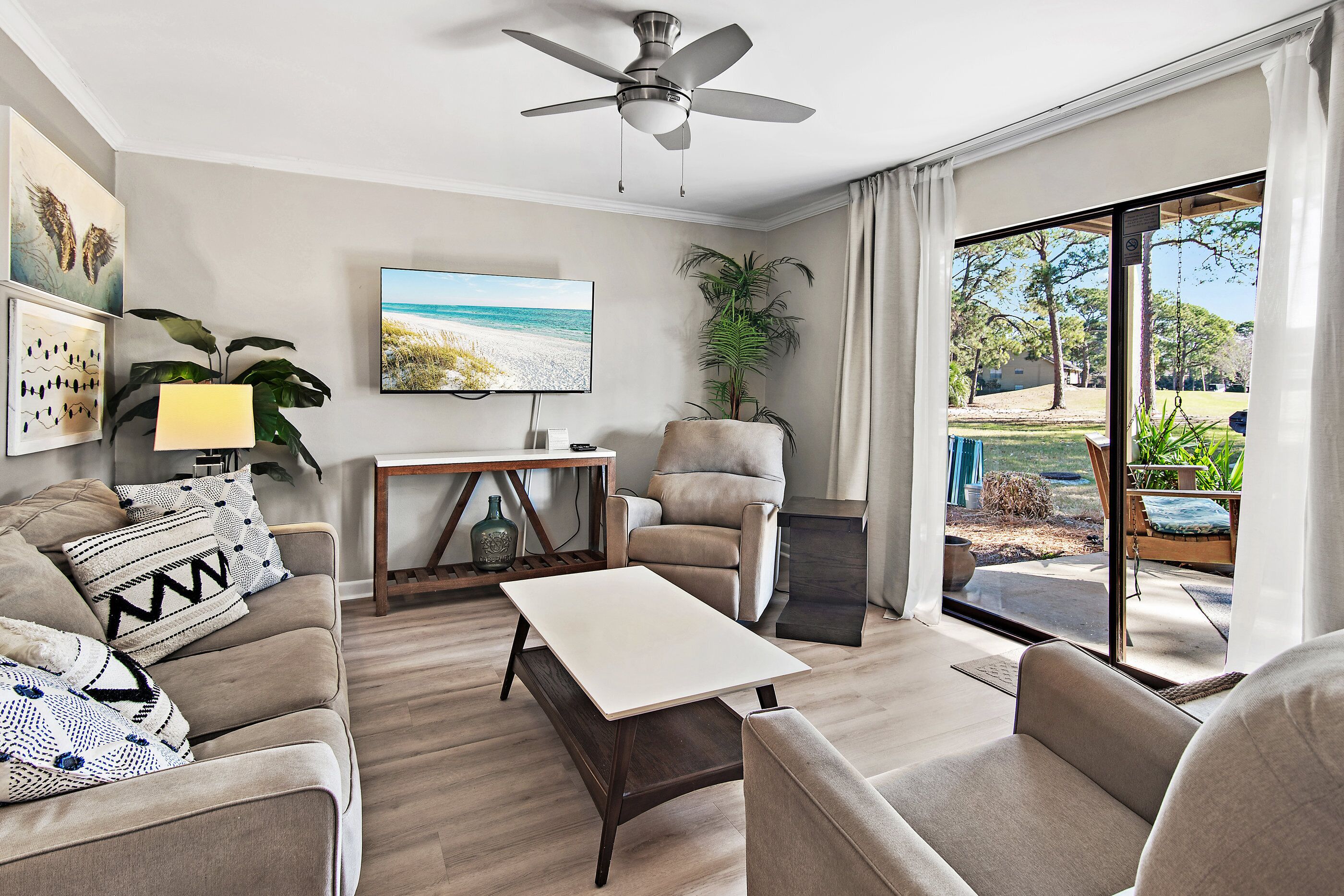 A living room with a sofa, armchair, coffee table, TV, ceiling fan, and a view of the outdoors.